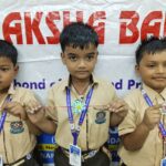 Three schoolchildren in beige uniforms pose with colorful circular crafts on strings in front of a banner at a school event wearing lanyards and badges